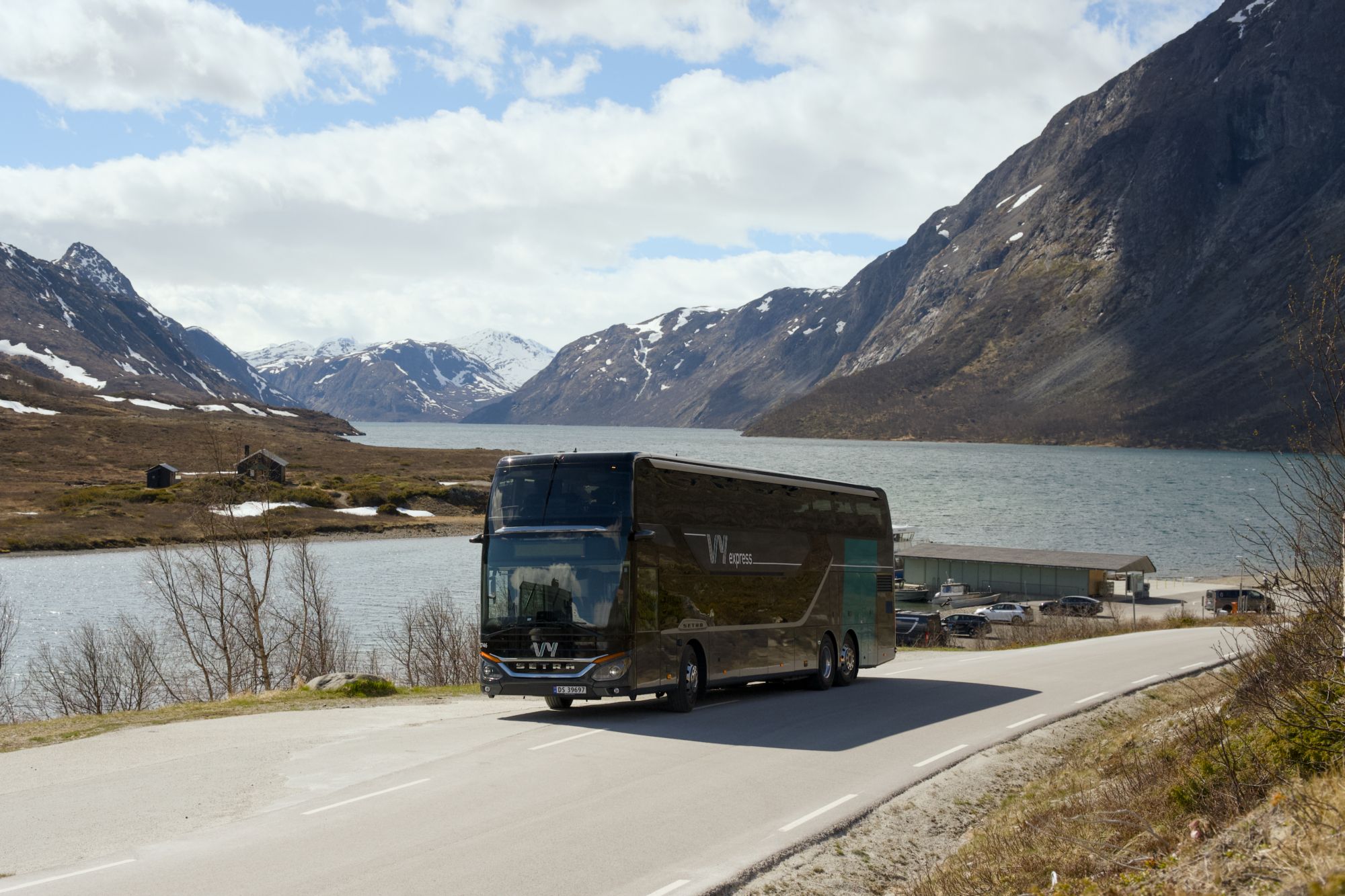 En Vy-buss på veien med fjord og fjell i bakgrunnen.