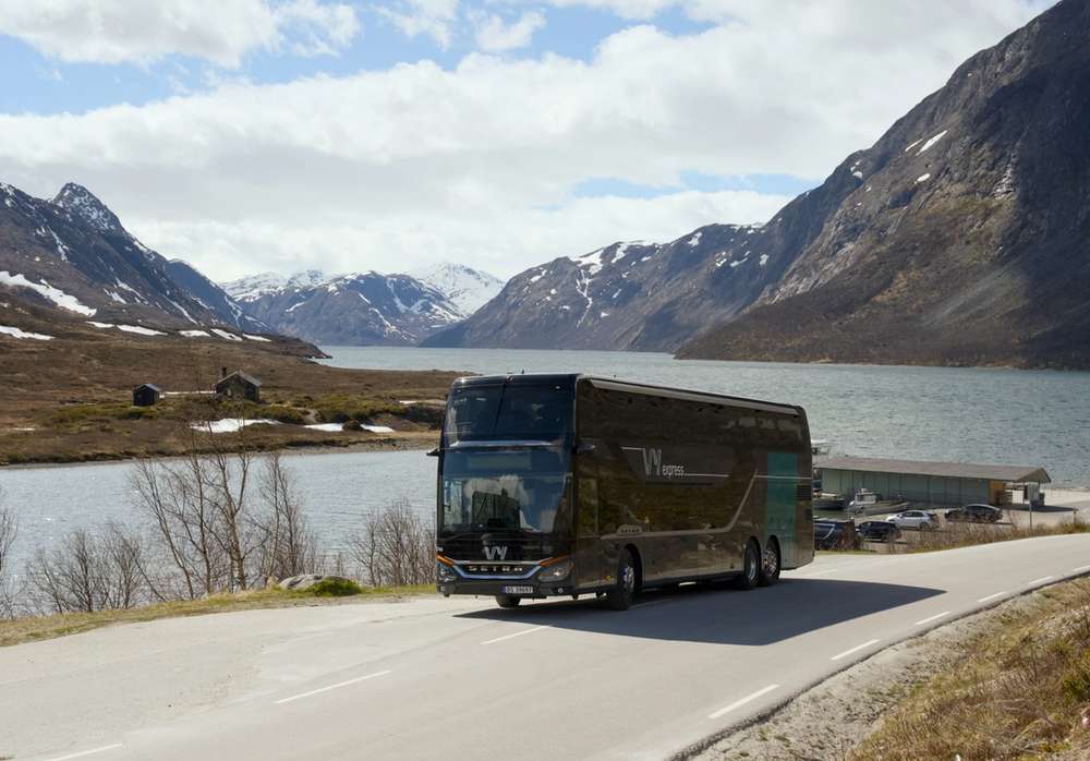 En Vy-buss på veien med fjord og fjell i bakgrunnen.