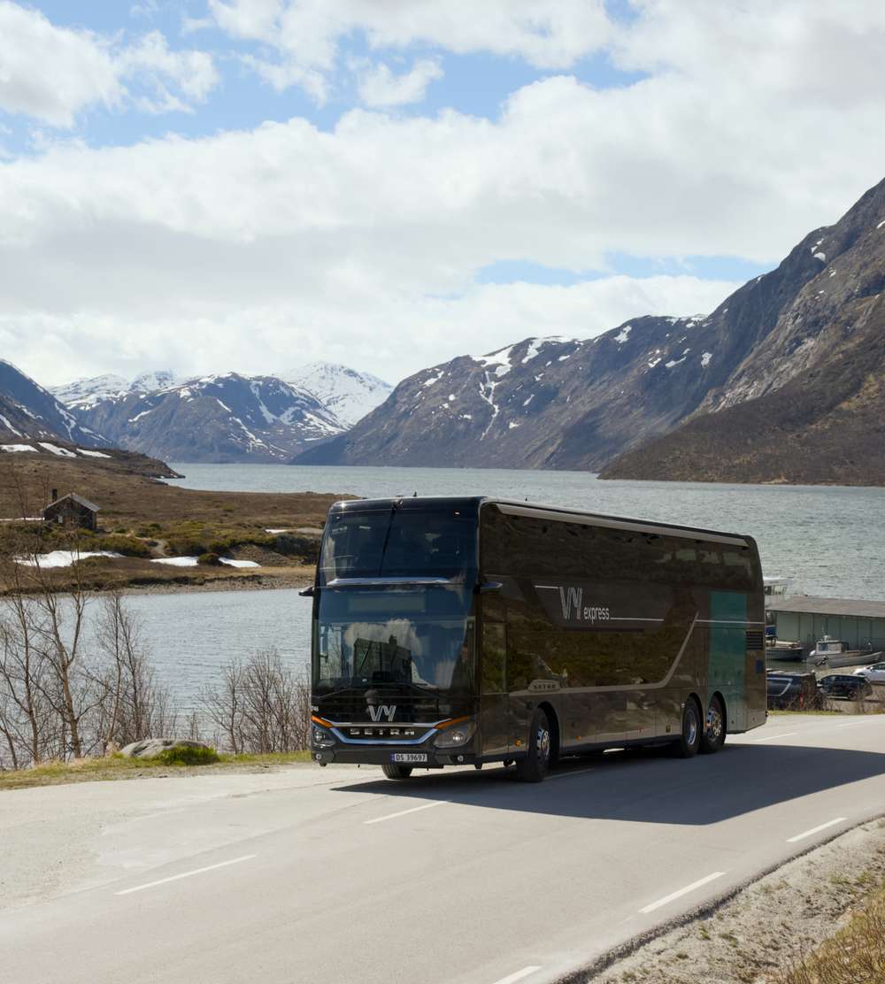 En Vy-buss på veien med fjord og fjell i bakgrunnen.