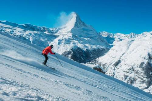 Person i rød jakke som står på ski nedover en bratt bakke med fjell.