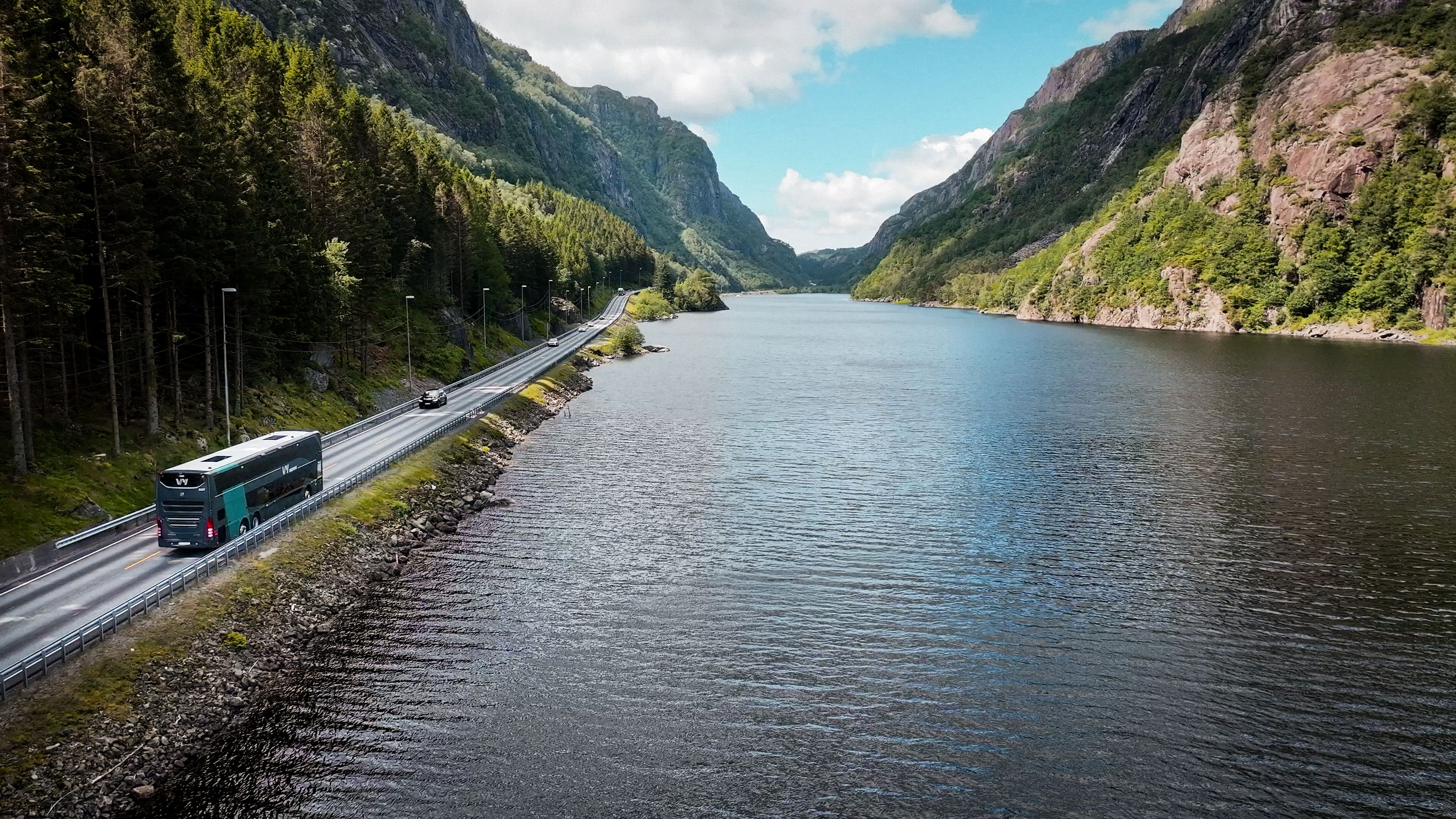En Vy-buss som kjører langs et vann med fjellandskap.