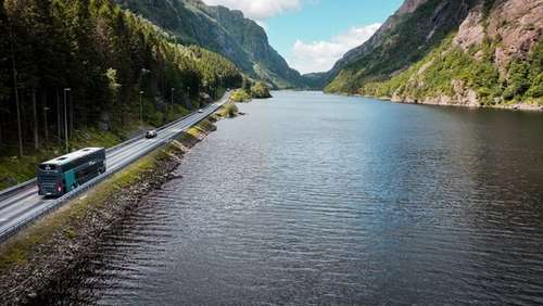 En Vy-buss som kjører langs et vann med fjellandskap.