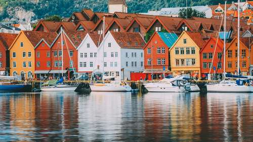 En rekke fargerike bygninger langs bryggen i Bergen, med båter på vannet og fjell bak.