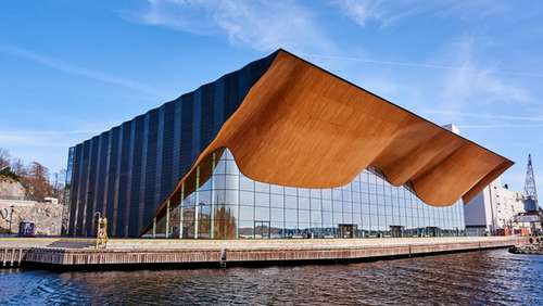 Kilden konserthus, en bygning av glass og treverk, med blå himmel over and vannet foran.
