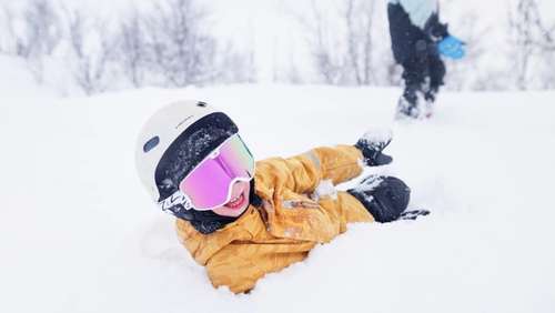 A child in a yellow winter suit and slalom helmet lies in the snow and smiles.