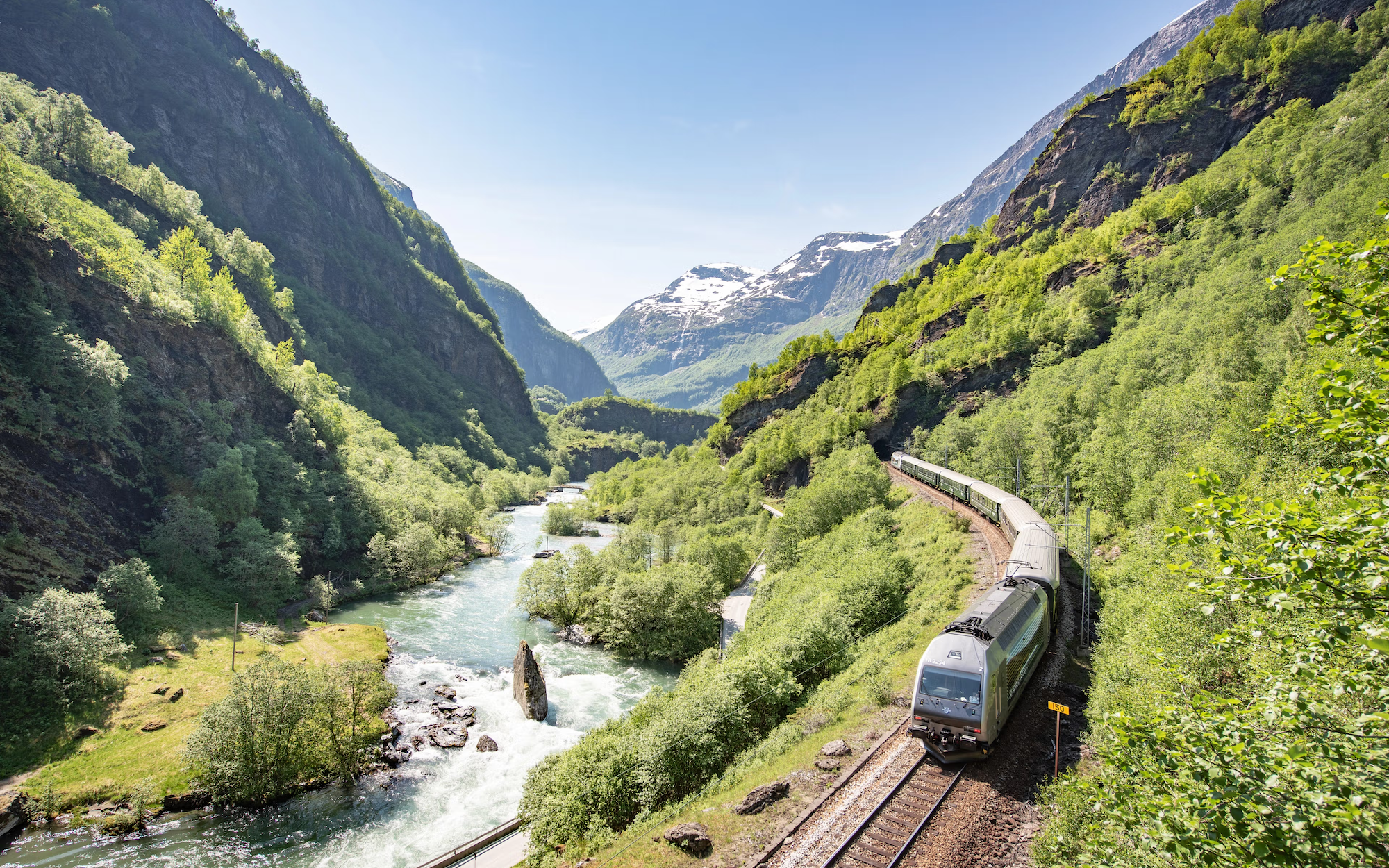 Tog som køyrer mellom høge fjell, ei elv og grøne omgivnader i fint vêr
