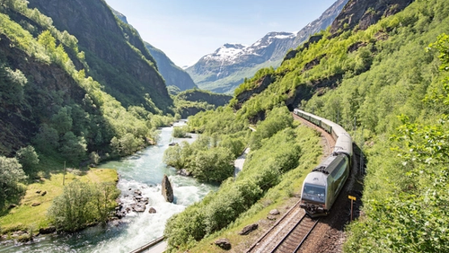 Tog som køyrer mellom høge fjell, ei elv og grøne omgivnader i fint vêr