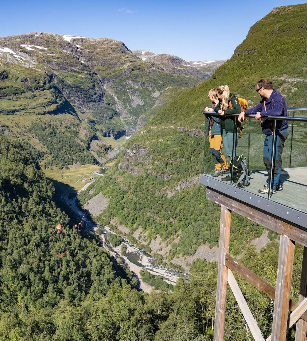 Zwei Personen stehen auf einer Aussichtsplattform mit Glasrand und blicken über das Flåmtal.