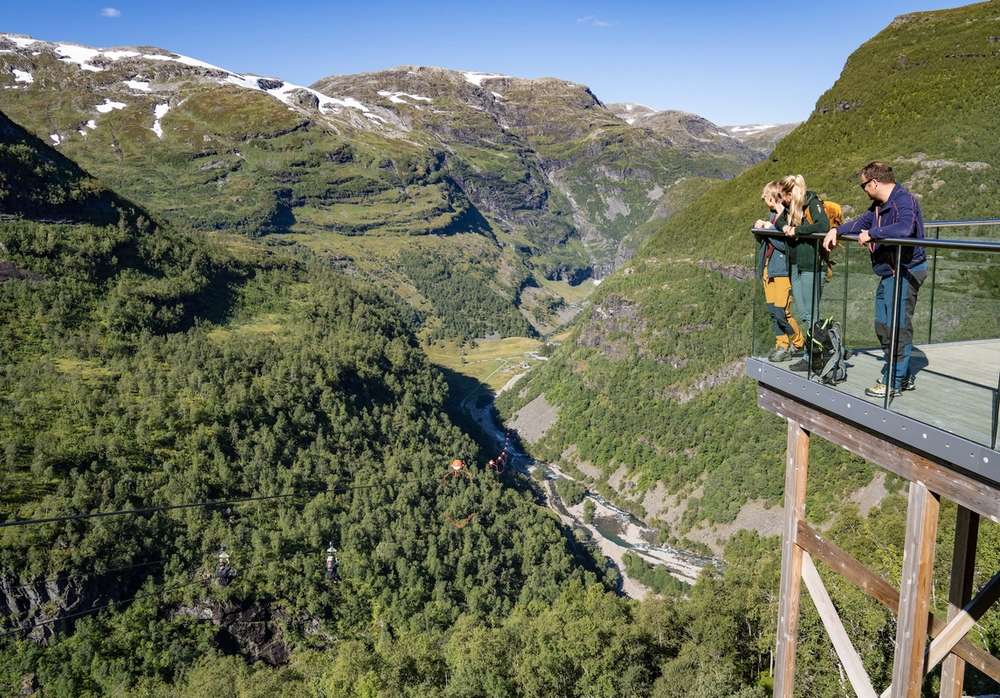 Zwei Personen stehen auf einer Aussichtsplattform mit Glasrand und blicken über das Flåmtal.