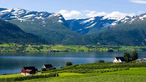 Bilde av snøbelagte fjell i bakgrunnen med grønne jorder og noen hus foran Gloppenfjorden i Sandane