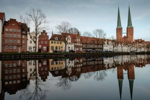 En rekke med hus langs en elv, bygningene reflekteres i vannet, to spir i bakgrunnen.