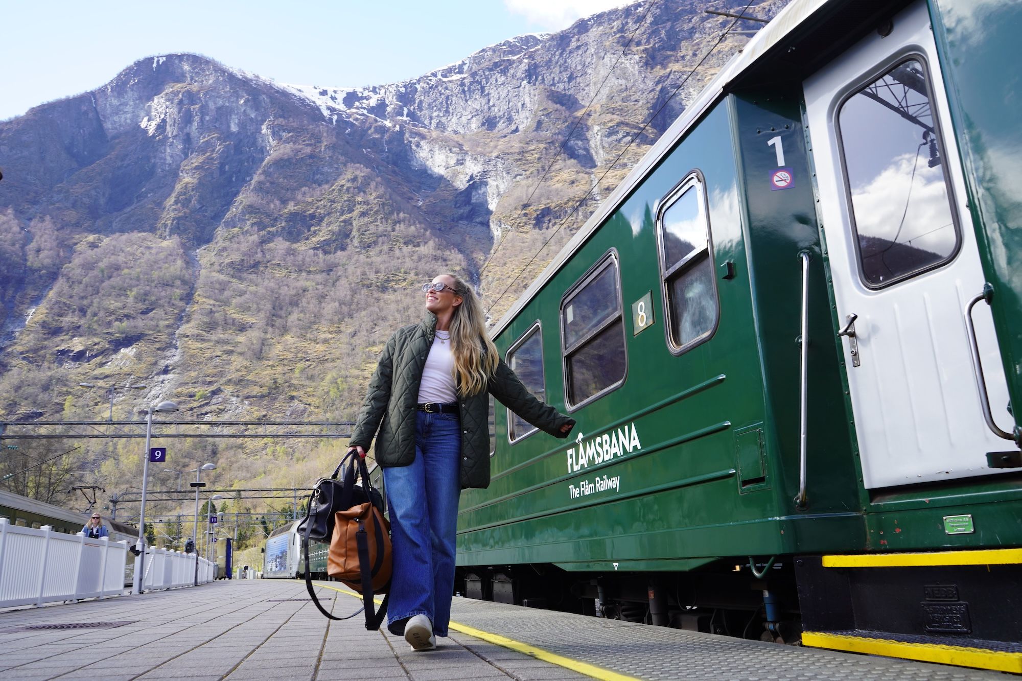 Woman walking past a green train, with mountains in the background.