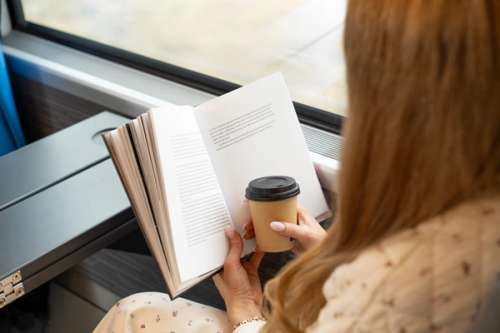Kvinne leser bok på toget med kaffe i handen.