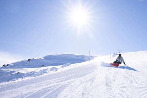 A person snowboards down a groomed slope with cloudless skies and sun.