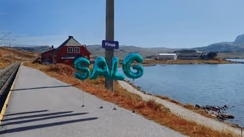 Vann med en vei som går langsmed, fjell og blå himmel i bakgrunnen. Et skilt hvor det står Finse, og Salg skrevet med ballonger.