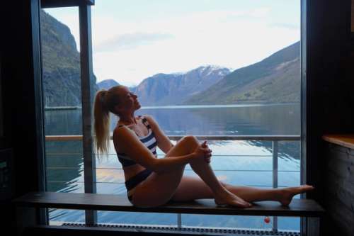 Woman sitting in front of a window in a sauna, a view of a lake and mountains.
