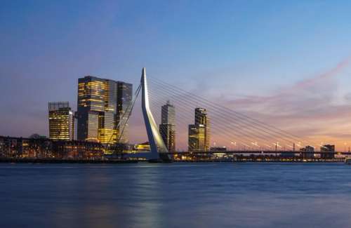 Kveldsbilde av Rotterdam, med høye bygninger, en bro og vann.