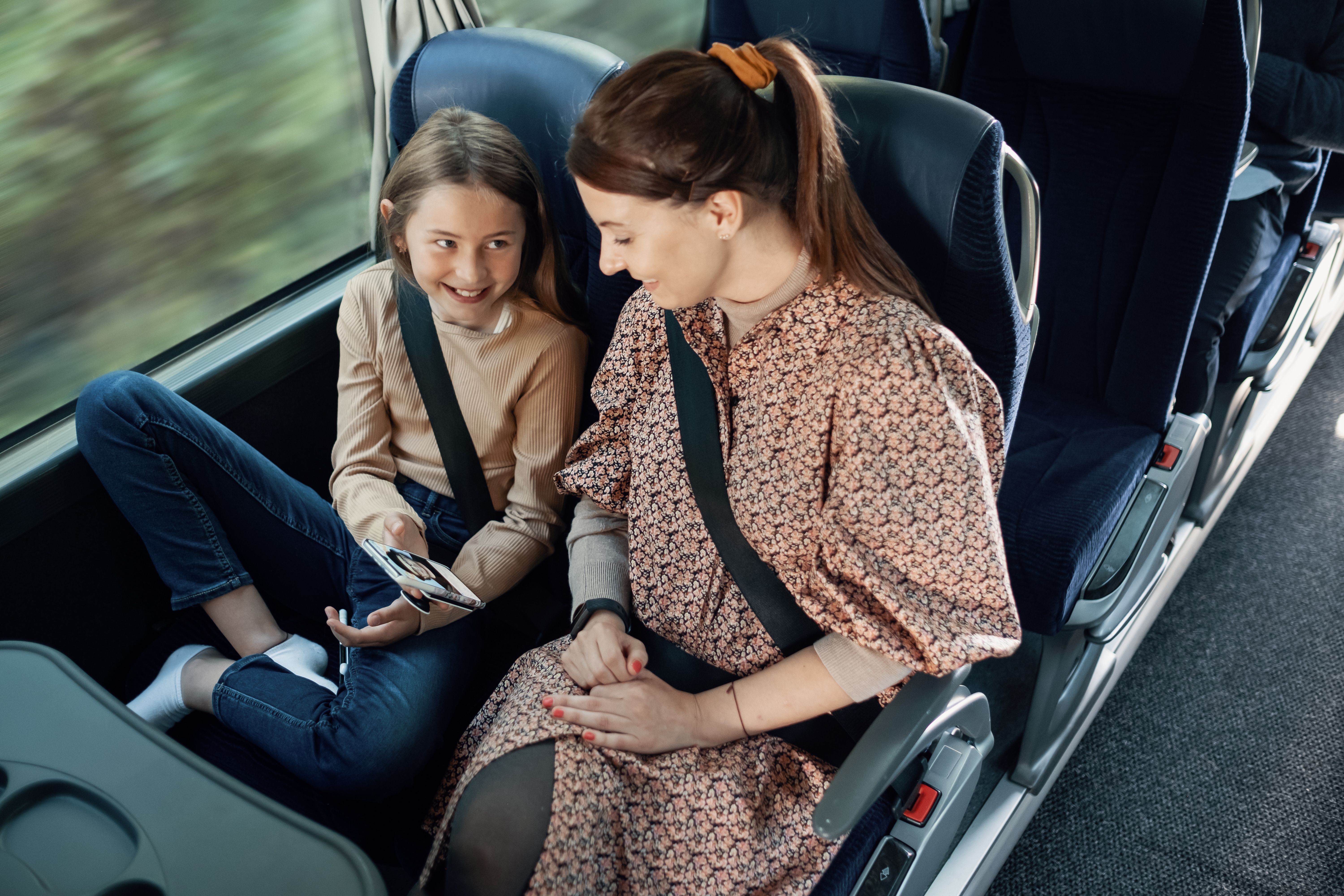 Datter viser noe på mobilen til moren sin mens de  sitter på bussen.