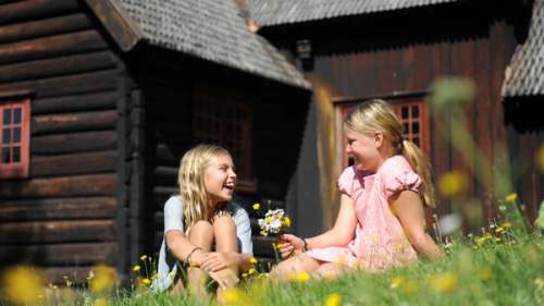 To jenter sitter i gresset foran en brun hytte.