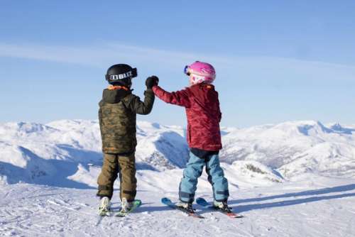 Two children high five while standing on slalom skis with a view of snow-covered mountains.