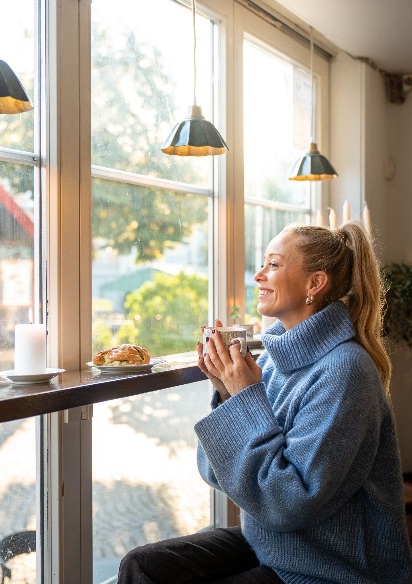 A woman with a blue sweater sitting with a coffee by a window looking out.