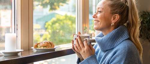 A woman with a blue sweater sitting with a coffee by a window looking out.