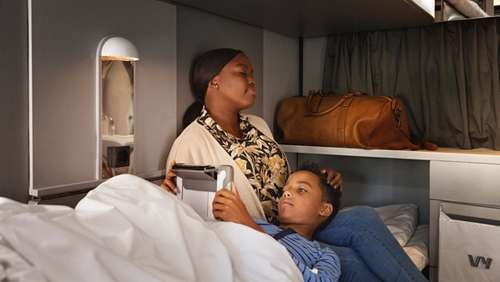 A woman sits in bed in a sleeping compartment on the Bergen Line train, with her son lying on her lap while he looks at an iPad.