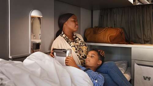 A woman sits in bed in a sleeping compartment on the Bergen Line train, with her son lying on her lap while he looks at an iPad.