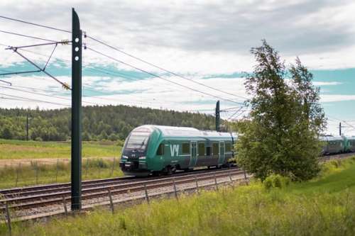 Et Vy-tog kjører på jernbaneskinnene med blå himmel og grønn natur rundt.