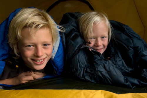 Gutt og jente smiler mot kamera, de ligger i soveposer i et telt.