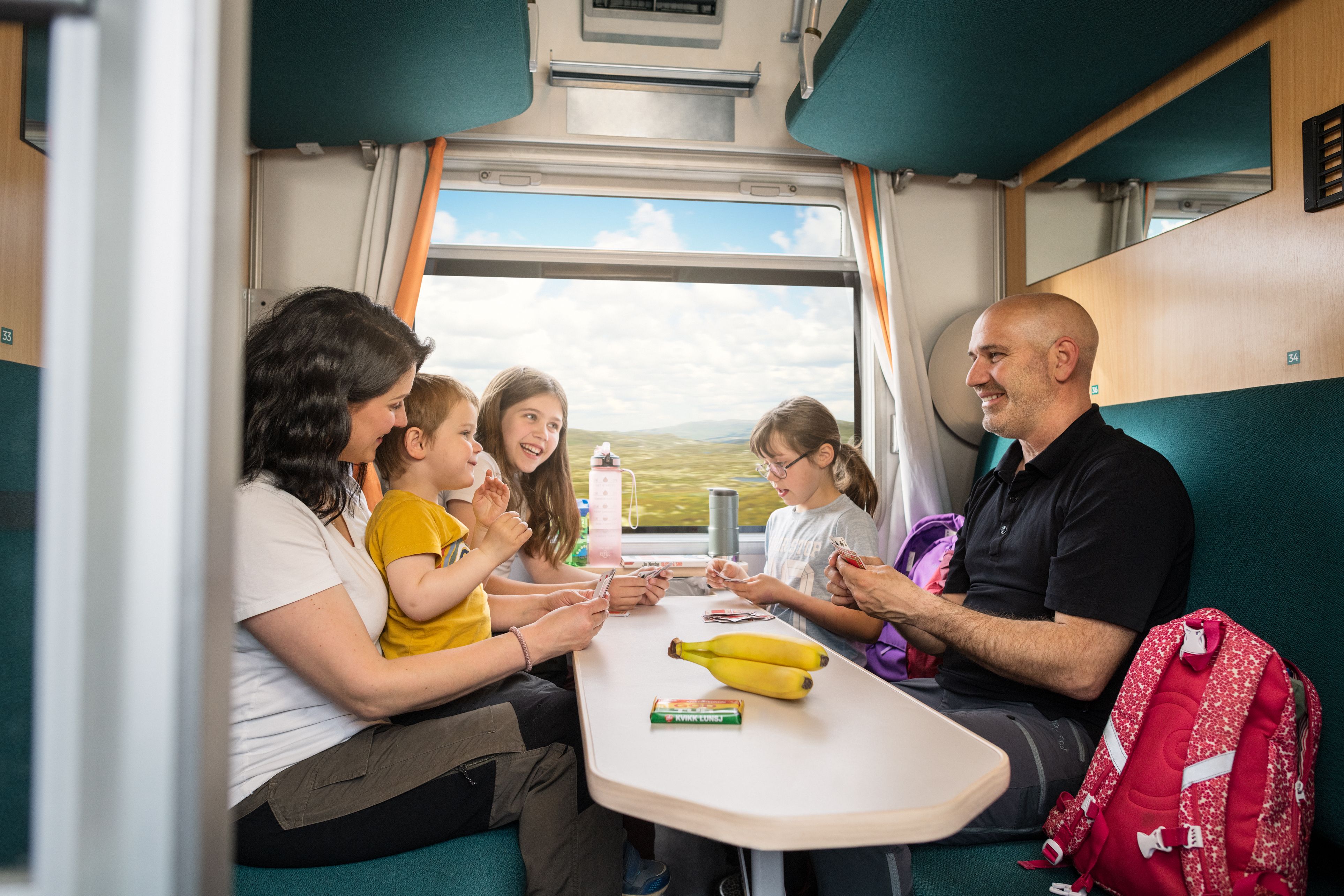 Innsiden av en kupé med en familie to voksne og tre barn som spiller kort ved et bord.