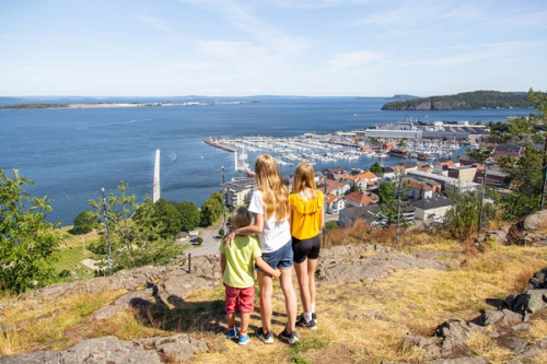 Tre barn står på en gressbakke og ser utover havna og sjøen ved en by.