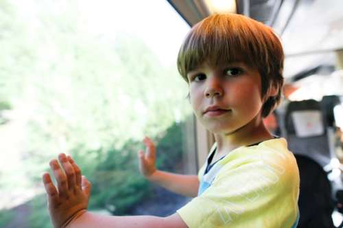 A boy wearing a yellow t-shirt looking a the camera. His hands are on the train window.