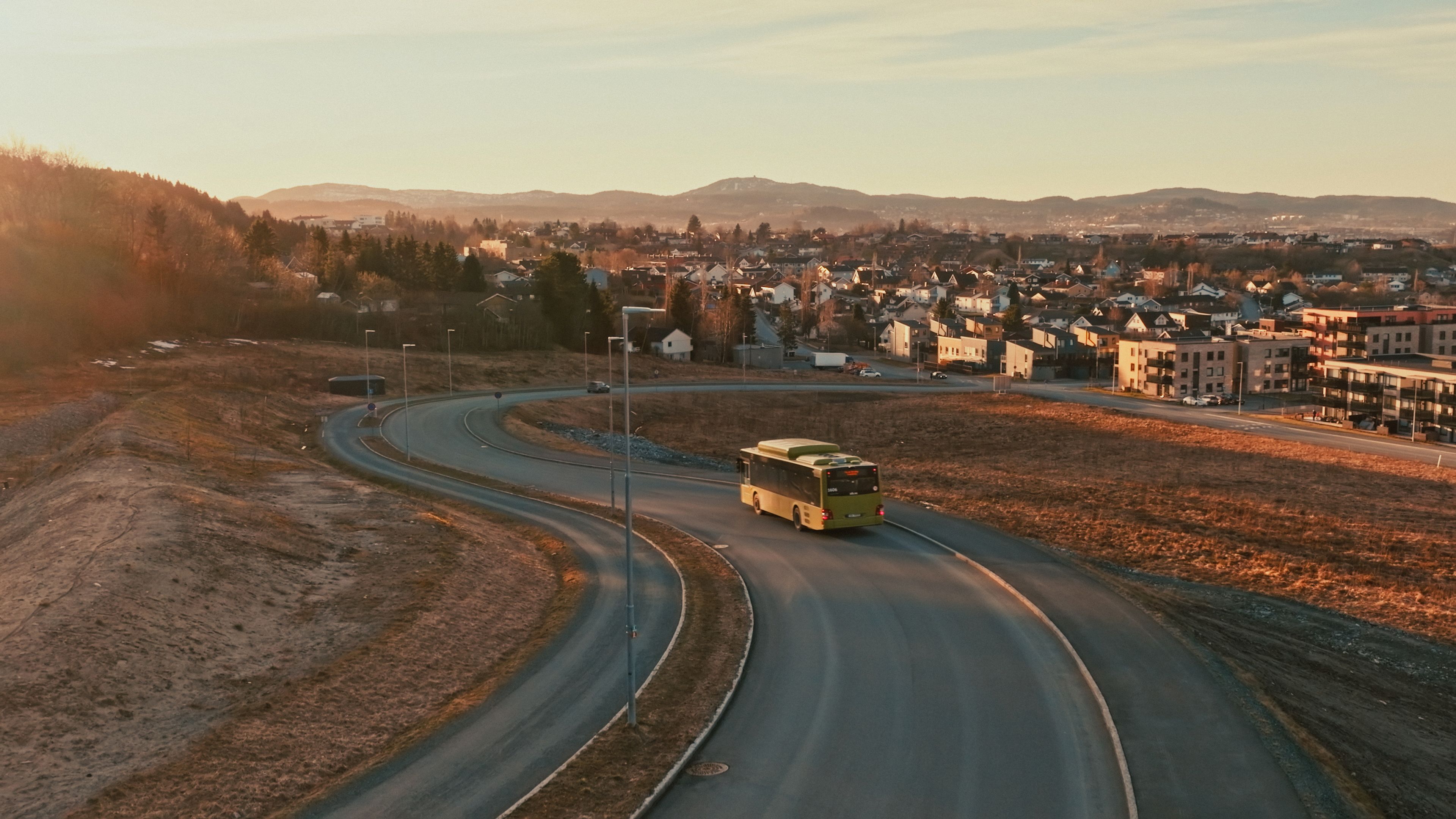 En grønn buss som kjører på veien med bebyggelse i bakgrunnen.