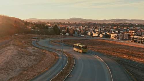 En grønn buss som kjører på veien med bebyggelse i bakgrunnen.