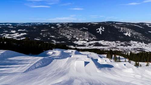 White ski slopes with jumps with blue sky and mountains in the background.