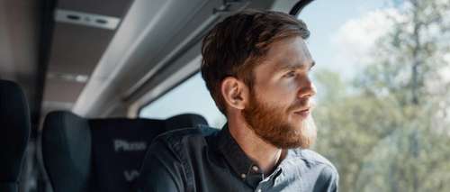 Man with a beard and shirt sits in a Pluss carriage on the Bergen Railway, looks out the window and enjoys a coffee.