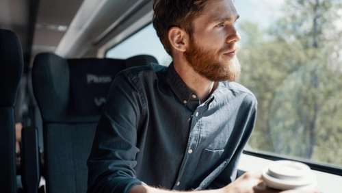 Mann med skjegg og skjorte sitter i Pluss-vogn på Bergensbanen, ser ut vinduet og nyter en kaffe.