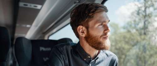 Mann med skjegg og skjorte sitter i Pluss-vogn på Bergensbanen, ser ut vinduet og nyter en kaffe.