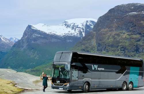 En dobbeldekker parkert med fjellandskap i bakgrunnen og en sjåfør som vinker ved siden av bussen.