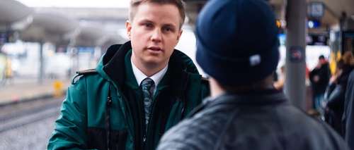 Konduktør Stian Fjellmyr Bye iført grønn Vy-jakke, står på en togstasjon og snakker med en passasjer