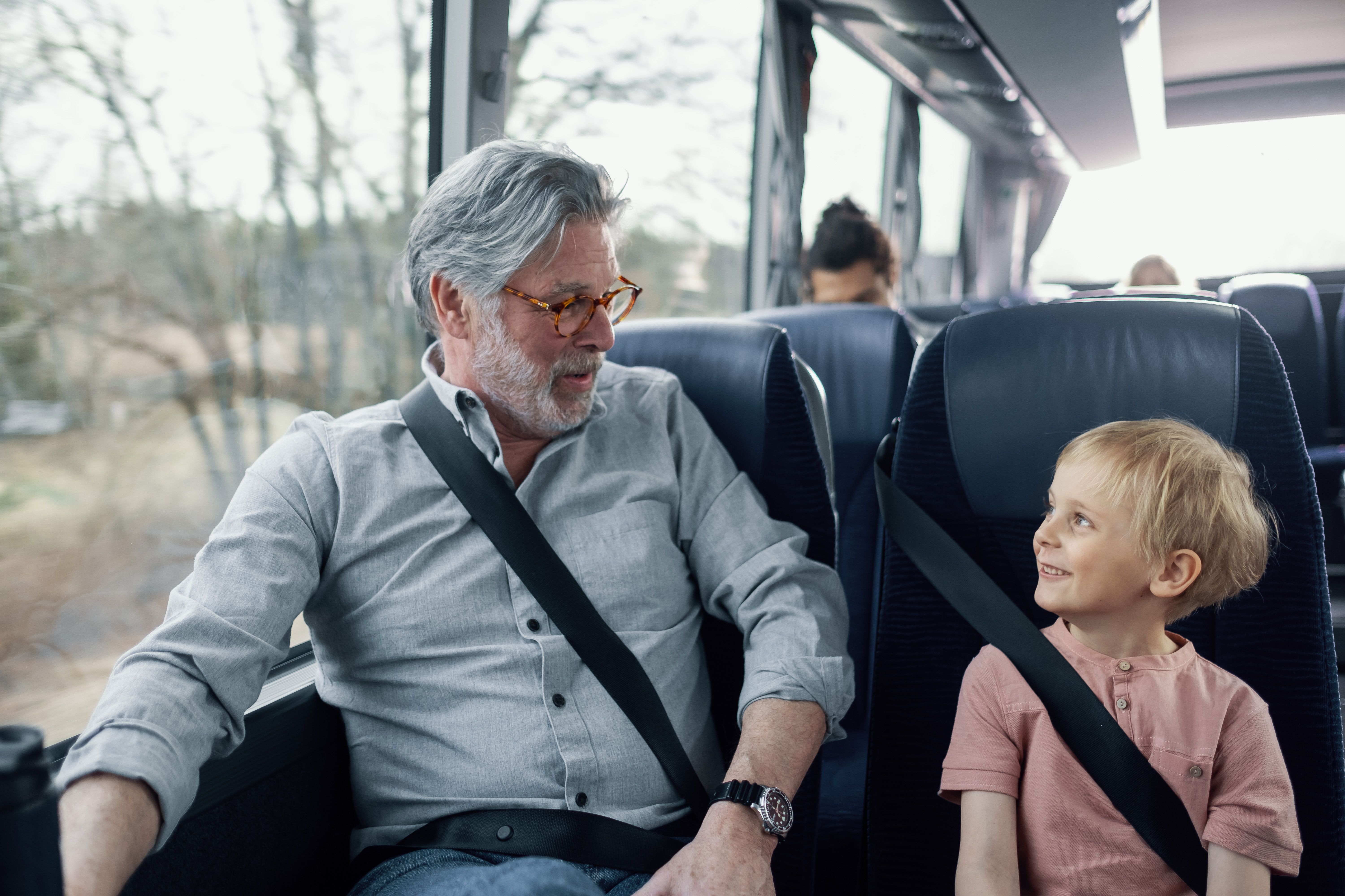 Eldre mann och ung gutt sitter vid varandra på bussen. De har på bilbelte och ser på varandra.