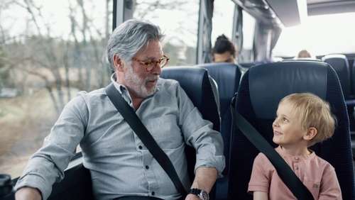 Eldre mann och ung gutt sitter vid varandra på bussen. De har på bilbelte och ser på varandra.