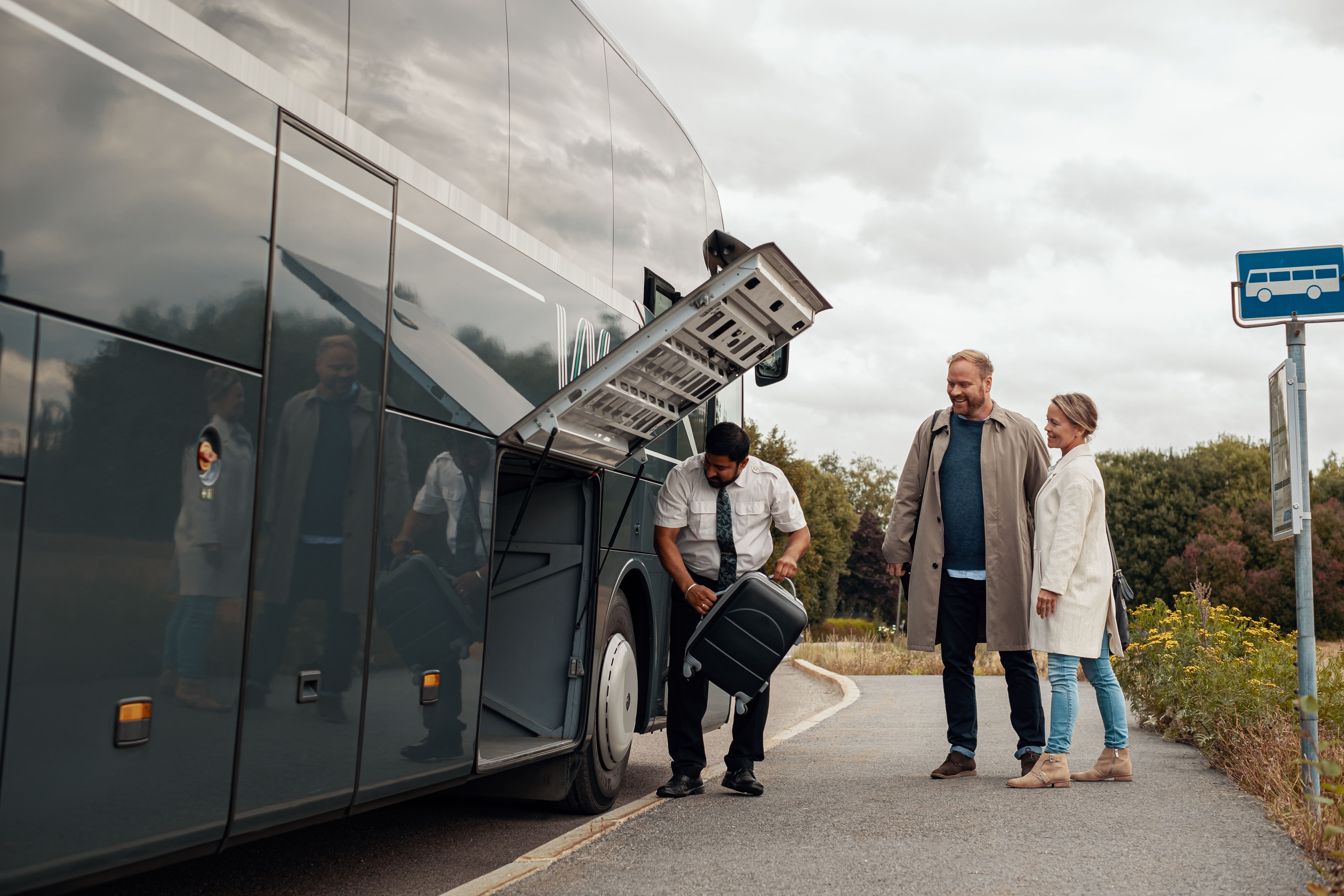 Busschaufför hjälper leende man och kvinna att lägga bagage i bussens bagageutrymme.