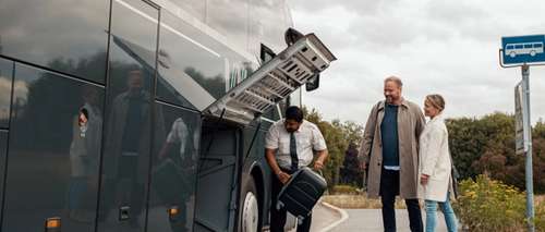 Busschaufför hjälper leende man och kvinna att lägga bagage i bussens bagageutrymme.
